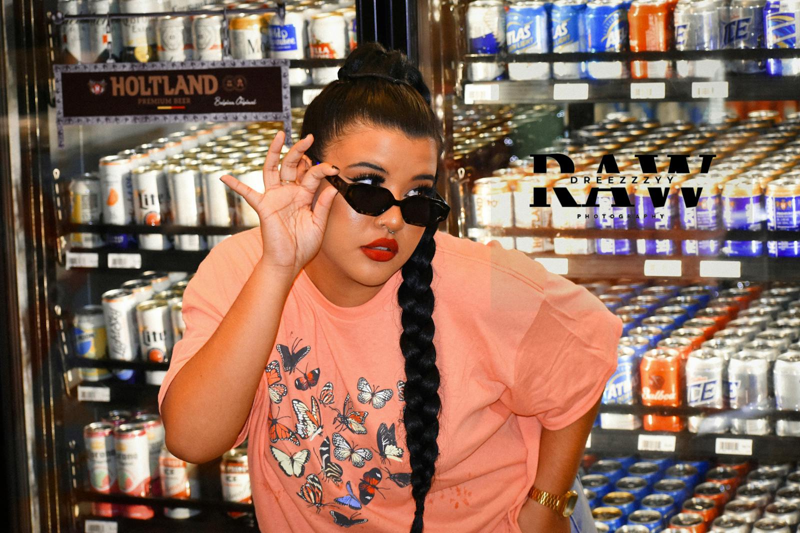 Fashionable woman in sunglasses and braids posing near beverage fridge indoors.